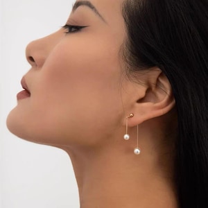 May include: A pair of gold dangle earrings with white pearl accents. The earrings feature a thin gold chain with a small pearl at the top and a larger pearl at the bottom.