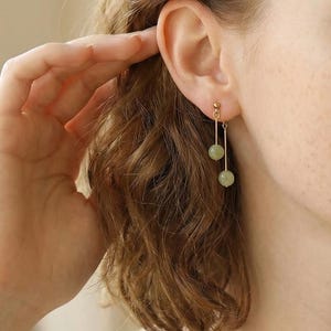Puede incluir: Pendientes dorados con un diseño de barra vertical, cada uno con una piedra preciosa redonda de color verde claro. Los pendientes se muestran en la oreja de una persona, con la mano tocando suavemente el cabello.