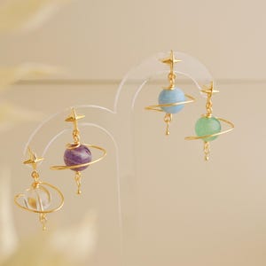 May include: Four gold-toned earrings shaped like planets with rings. Each earring features a small star-shaped stud at the top and a spherical gemstone bead in clear, purple, light blue, or green, encircled by a thin gold wire ring.