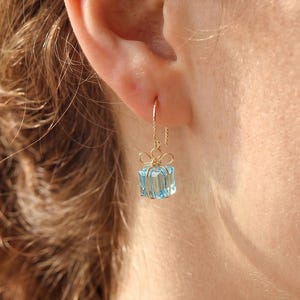 May include: A gold earring featuring a light blue, cube-shaped gemstone. The gemstone hangs from a fine gold wire and is decorated with a small bow. The earring is displayed on a person's ear.
