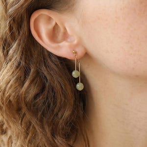 May include: A gold-toned drop earring featuring two small, smooth, pale green spherical beads suspended at different lengths from a thin metal wire. The earring is attached to the earlobe with a small gold-toned stud post and back closure.