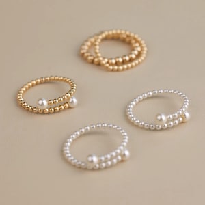 Puede incluir: Se muestran cuatro anillos sobre una superficie neutra. Dos son dorados, uno con un diseño en espiral y dos perlas, y el otro una banda simple. Los otros dos anillos son plateados, con un diseño en espiral y perlas.