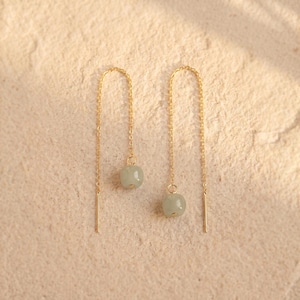 May include: A pair of delicate gold-colored chain earrings. Each earring features a small, pale green, rounded bead suspended from a fine chain. A straight, gold-colored bar hangs at the bottom of each chain. The earrings are displayed on a light beige surface.