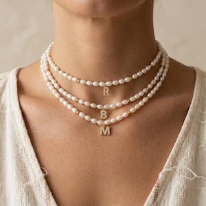 May include: Three-strand pearl necklace with gold accents and initial pendants. Each necklace features a delicate string of white pearls, interspersed with small gold beads. The pendants are gold and encrusted with small crystals, displaying the letters R, B, and M.