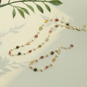 May include: A delicate gold chain necklace and bracelet with small, colourful gemstones and white pearls. The necklace and bracelet are both made of gold chain and feature a mix of pink, green, and brown gemstones.