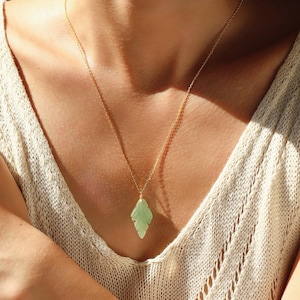 May include: A gold-colored necklace with a light green leaf-shaped pendant. The pendant is a pale green color and hangs from a delicate gold chain. The necklace is worn against a light-colored top.