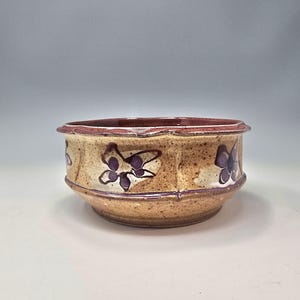 May include: A round ceramic bowl with a speckled tan and beige glaze, featuring a dark purple floral pattern around the exterior. The rim and base are accented with a deep maroon band, and the bowl has a slightly textured, hand-formed appearance.