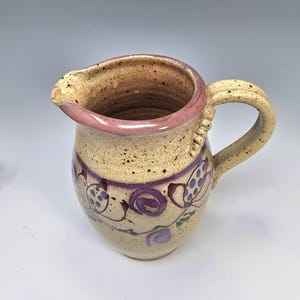 May include: A handmade ceramic pitcher with a speckled beige body, a pink rim, and a curved handle. The pitcher is decorated with purple floral designs and green leaves. The handle has a textured design.