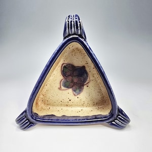 May include: A triangular ceramic dish with a blue rim and accents. The interior is beige with a floral design in the center. The dish has three small feet and a handle on one side.