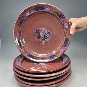 May include: A stack of six ceramic plates. The plates are a reddish-brown color with a floral design in blue, purple, and green. The plates have a glossy finish and are stacked on top of each other.