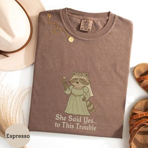 May include: Espresso-colored t-shirt with a raccoon graphic wearing a veil and holding a diamond ring. The text "She Said Yes... to This Trouble" is printed below the graphic. A gold necklace and sandals are also visible.