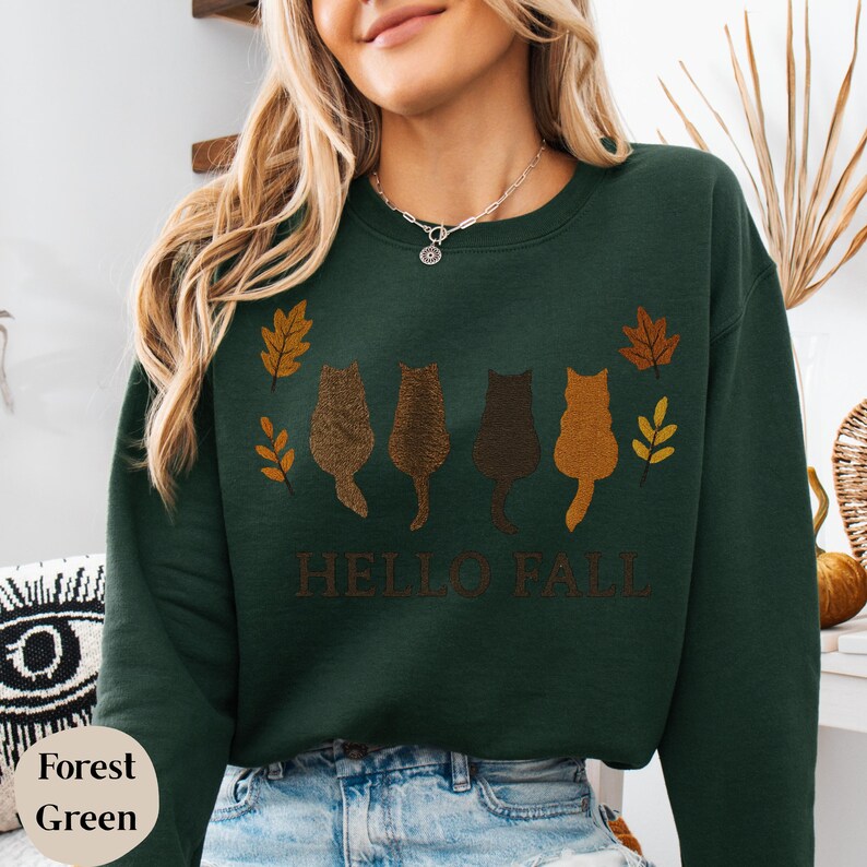 May include: Forest green sweatshirt with a fall-themed design. The design features four cat silhouettes in shades of brown, with autumn leaves and the text "HELLO FALL" in brown. The sweatshirt is made of a soft material.