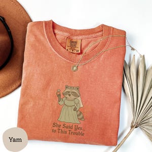 May include: A coral-colored t-shirt with a raccoon bride graphic holding a ring. The text "She Said Yes... to This Trouble" is below the graphic. A gold necklace and a brown hat are also visible.