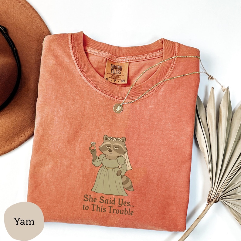 May include: A coral-colored t-shirt with a raccoon bride graphic holding a ring. The text "She Said Yes... to This Trouble" is below the graphic. A gold necklace and a brown hat are also visible.