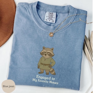 May include: A blue jean Comfort Colors t-shirt featuring a cartoon raccoon wearing a green tunic and the text "Engaged to My Favorite Menace." A gold necklace with a pendant is also visible.