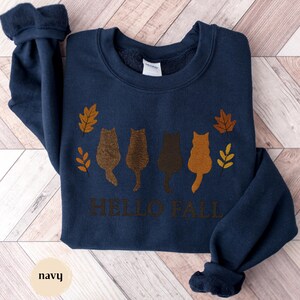 May include: Navy blue sweatshirt featuring four cat silhouettes in shades of brown, with fall leaves and the text "HELLO FALL" in brown. The word "navy" is at the bottom left. A cozy, long-sleeved top.