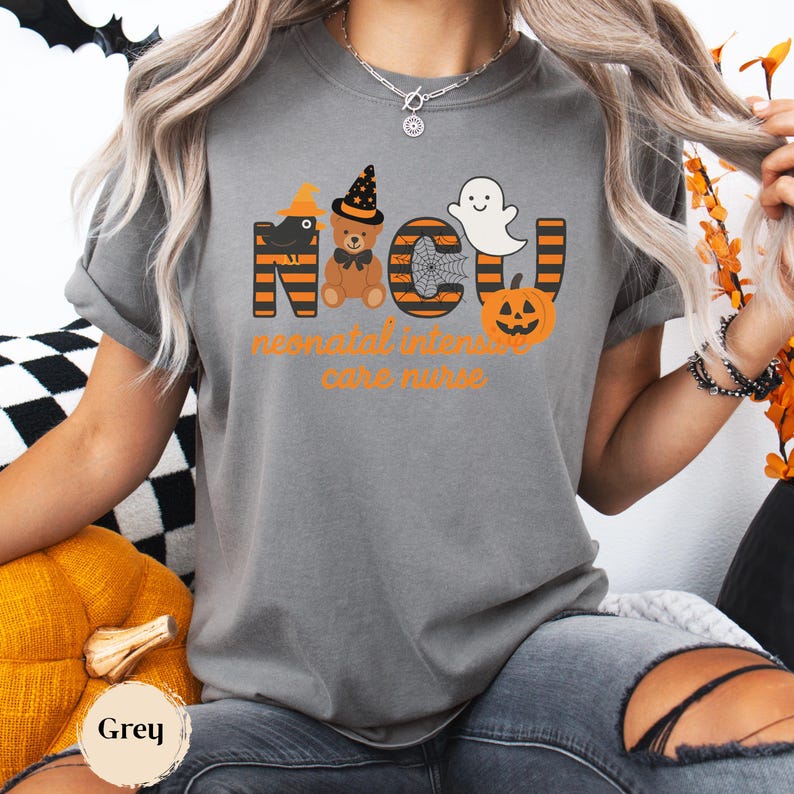 Pu&ograve; includere: T-shirt grigia con un design a tema Halloween. Il design include le lettere "NICU" con un corvo, un orsacchiotto, una ragnatela, un fantasma e una zucca. Il testo "neonatal intensive care nurse" &egrave; sotto.