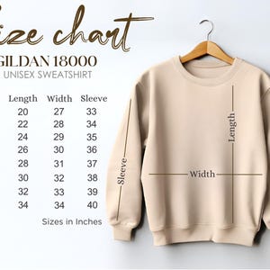 May include: A beige Gildan 18000 unisex sweatshirt hangs on a wooden hanger. A size chart is displayed, showing measurements in inches for length, width, and sleeve for sizes S to 5XL. The text "Size chart" is at the top.
