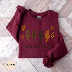 May include: Maroon sweatshirt with the text "HELLO FALL" and four cat silhouettes in shades of brown and orange. The design includes fall leaves. The word "maroon" is at the bottom left.