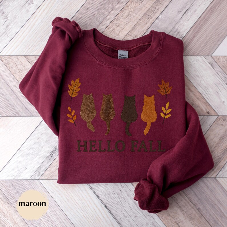 May include: Maroon sweatshirt with the text "HELLO FALL" and four cat silhouettes in shades of brown and orange. The design includes fall leaves. The word "maroon" is at the bottom left.