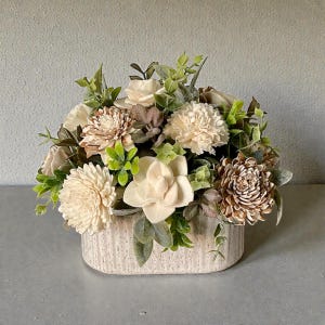 May include: A floral arrangement featuring a variety of artificial flowers in shades of cream, brown, and green. The flowers are arranged in a rectangular, textured, off-white vase. The arrangement includes various flower types and greenery.