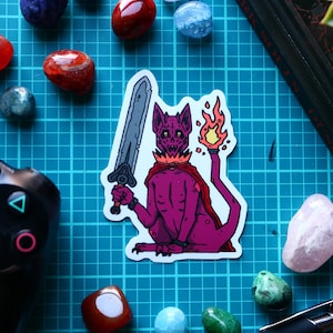 May include: A sticker depicting a purple creature holding a sword and a flaming torch. The creature wears a cape, set against a blue grid. There are colourful stones and a game controller. The sticker is a fantasy-themed design.