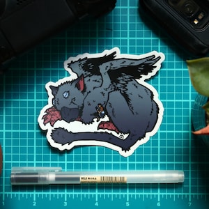 May include: A gray and black winged cat sticker with a red and white design. The sticker is on a blue cutting mat.