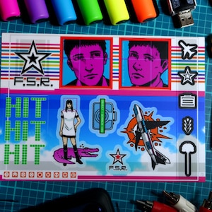Może przedstawiać: Arkusz naklejek z grafiką w stylu pop-art. Naklejki przedstawiają portrety, kobietę na krokodylu, rakietę i napis "HIT". Inne elementy to długopisy, markery i kontroler do gier.