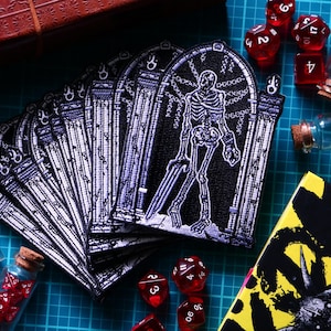 May include: A collection of embroidered patches featuring a white skeleton standing within a gothic archway on a black background. The patches are arranged in a fan shape alongside several small glass vials filled with red polyhedral dice.