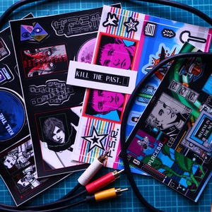 May include: A collection of colorful stickers with various designs, including portraits, geometric shapes, and the text "KILL THE PAST." The stickers are arranged with audio cables and a handheld gaming device.