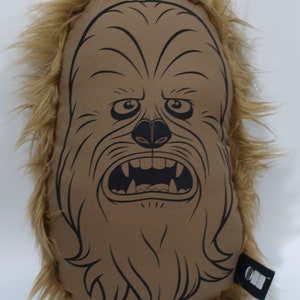 Puede incluir: Una almohada de felpa marrón con una ilustración en blanco y negro de la cara de Chewbacca. La almohada tiene un borde de piel marrón.