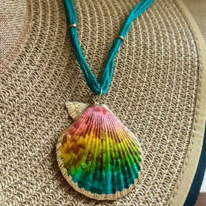 May include: A colorful seashell pendant necklace. The pendant is painted with a rainbow gradient of pink, yellow, green, and blue. The shell is outlined in gold and hangs from a teal cord necklace. The necklace is resting on a woven straw hat.