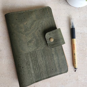 May include: A green cork notebook with a flap closure and a gold button. The notebook is lying on a light beige surface with a pen next to it.