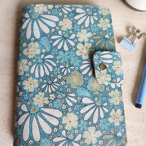 May include: A floral-patterned journal with a light blue background and white and tan daisy-like flowers. The journal has a flap closure with a metal snap. A small plant, a paper clip, and a blue pen are in the background.