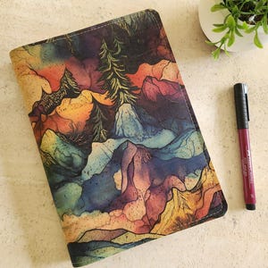 Puede incluir: Un diario cerrado con un diseño de paisaje de montaña colorido. La portada presenta una ilustración estilo acuarela con tonos de azul, naranja, amarillo y verde. Un bolígrafo burdeos descansa junto al diario, y una pequeña planta en maceta está en el fondo.
