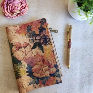 May include: A cork zipper pouch with a floral design featuring pink, orange, and blue flowers. A matching pen with gold accents and a pink floral pattern sits next to the pouch. A pink flower and a small potted plant are also in the image.