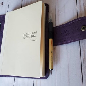 May include: A purple cork notebook cover with a cream-colored Hobonichi Techo 2022 Cousin planner inside. A black pen with Japanese text is clipped to the cover. Decorative bookmarks are visible in a pocket.