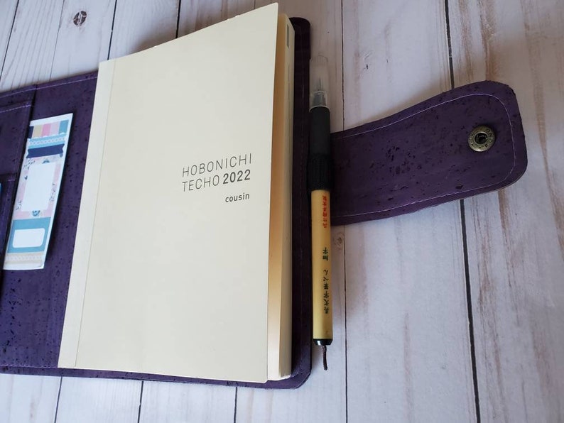 May include: A purple cork notebook cover with a cream-colored Hobonichi Techo 2022 Cousin planner inside. A black pen with Japanese text is clipped to the cover. Decorative bookmarks are visible in a pocket.