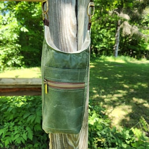 May include: A green waxed canvas crossbody bag with a brown zip and a small front pocket. The bag has a cream interior and is suspended by a braided strap with brass-coloured hardware.