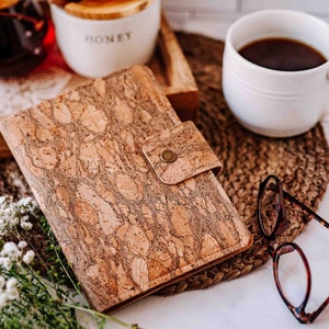 May include: A cork notebook with a button closure sits on a woven mat, next to a white mug of coffee and a pair of tortoiseshell glasses. A wooden tray with a jar of honey and a glass of liquid is in the background.