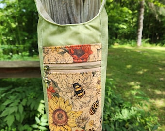 Puede incluir: Un bolso bandolera verde con un diseño floral y de abejas. El bolso tiene un bolsillo frontal de corcho con girasoles, abejas y flores rojas. Tiene cierre de cremallera y una correa de cadena.
