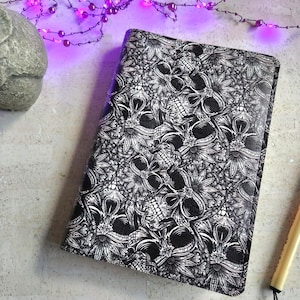 May include: A black and white journal with a skull and floral pattern. The journal is rectangular and has a decorative design. A black and gold pen is next to the journal. A stone owl statue is in the background.