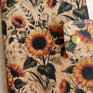 May include: A cork-like material notebook cover with a sunflower design. The cover features a repeating pattern of sunflowers in shades of yellow, orange, and brown, with green leaves and stems. A flap with a brass button closure is visible.