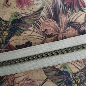 May include: Close-up of a fabric with a hummingbird and floral print. A light gray zipper with a dark metal zipper pull is placed diagonally across the fabric. The fabric has a vintage, botanical theme with green, yellow, and pink hues.