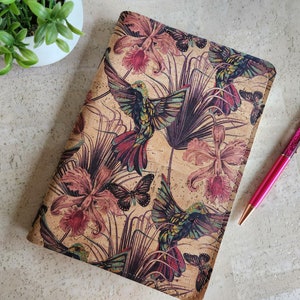 Puede incluir: Cubierta de cuaderno de corcho con un diseño floral y de colibrí. La cubierta presenta colibríes rosas y verdes con detalles florales sobre un fondo de corcho.