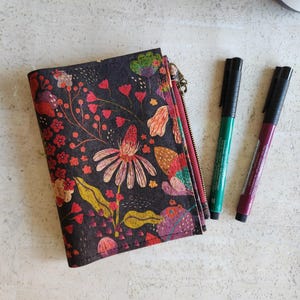 May include: A black floral wallet with a red zipper, featuring a colorful botanical design with red, pink, and yellow flowers. Two pens, one green and one burgundy, are placed next to the wallet. The wallet is closed.