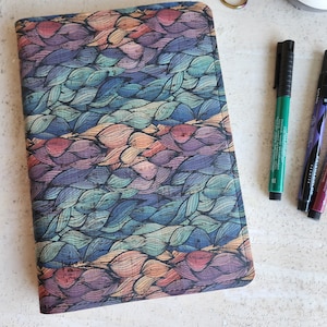 May include: A rectangular journal with a colorful, textured cover featuring a repeating leaf-like pattern in shades of blue, purple, orange, and green. The journal is next to a pair of small scissors and several markers.
