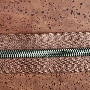 May include: Close-up of a brown zipper with a metallic green coil. The zipper tape is a light brown color with a textured, woven pattern. The zipper is centered on a brown, textured surface.