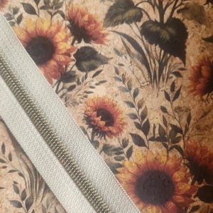May include: Close-up of a fabric with a sunflower pattern in shades of yellow, orange, and brown. A light gray zipper is diagonally across the image. The zipper teeth are silver.