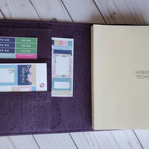 May include: Open purple cork planner with a cream-colored Hobonichi Techo 2022 cousin notebook. The planner has pockets holding colorful sticky notes with "To Do" and "Happy Birthday" labels, and floral designs. The planner is open.